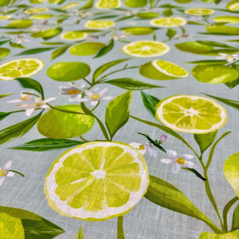May include: A light blue fabric with a repeating pattern of green limes, lime slices, white flowers, and green leaves. The limes are whole and sliced, with a detailed illustration of the fruit.