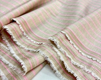 Könnte beinhalten: Beigefarbener Stoff mit rosa Streifen und Fransen. Der Stoff ist gefaltet und zeigt die Textur und das Muster. Die rosa Streifen sind gleichmäßig auf dem beigen Hintergrund verteilt und erzeugen einen klassischen Look. Der Fransenrand verleiht einen Hauch von rustikalem Charme.
