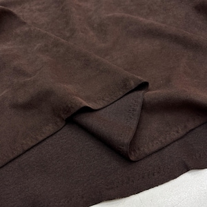 May include: Close-up of a folded piece of brown fabric. The fabric has a soft, textured appearance with visible folds and shadows, creating depth. The color is a rich, dark brown.