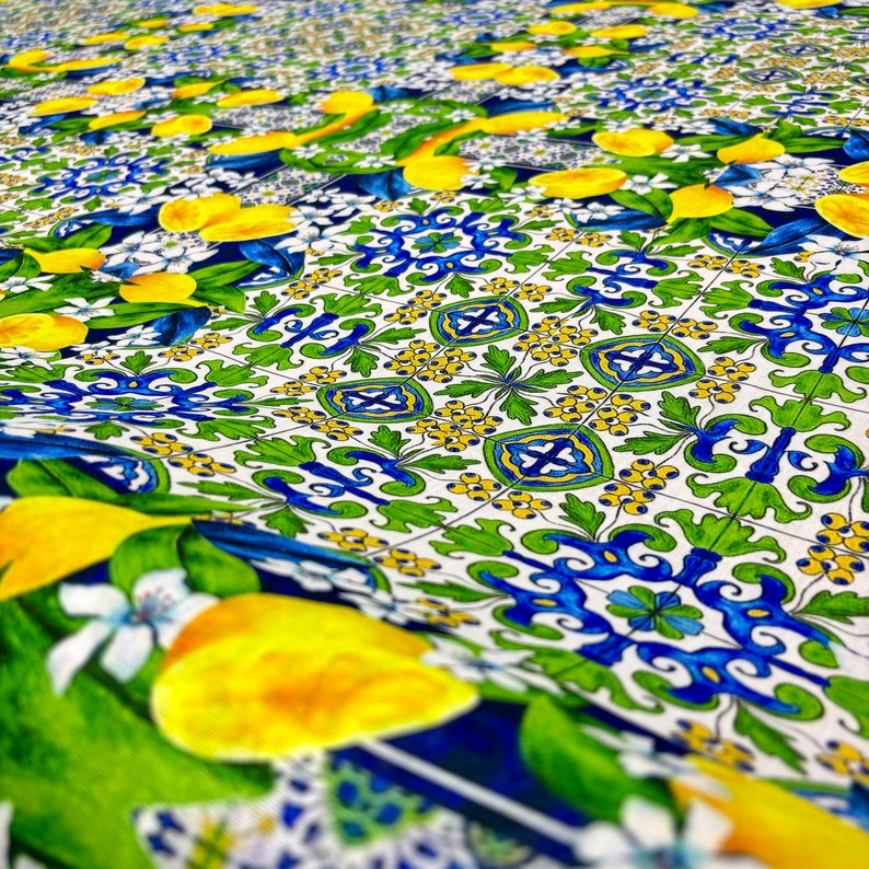 May include: A white fabric with a repeating pattern of blue, green, and yellow lemons and floral designs. The fabric has a textured surface and a slightly wrinkled appearance.