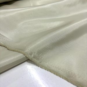 May include: Close-up of a pale green fabric with a silky sheen. The fabric is folded, showing the texture and the raw edge of the material. The color is a soft, muted green, suitable for apparel or home decor.