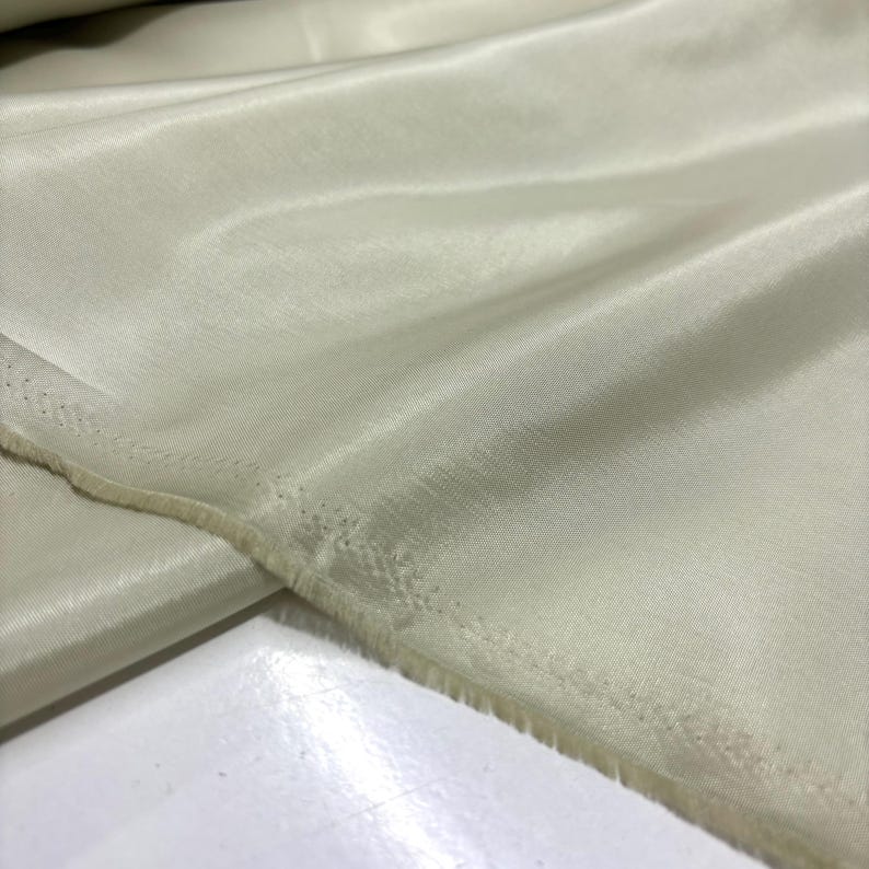 May include: Close-up of a pale green fabric with a silky sheen. The fabric is folded, showing the texture and the raw edge of the material. The color is a soft, muted green, suitable for apparel or home decor.