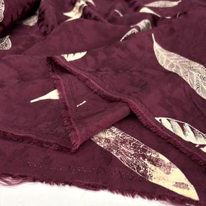 May include: Burgundy fabric featuring a white feather pattern. The material has a textured surface and a fringed edge. The feathers vary in size and design, creating a visually interesting textile.