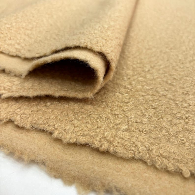 May include: Close-up of a beige curly fleece fabric. The fabric is folded over itself, showing the texture of the fleece.