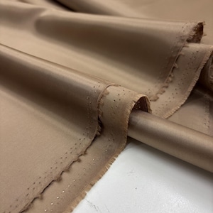May include: A bolt of solid tan fabric with a smooth, satin-like finish and a subtle diagonal twill weave. The material features a finished edge with a row of small, evenly spaced pinholes running along the length of the selvage border.