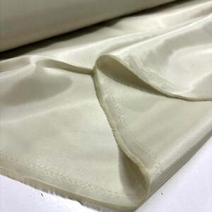 May include: A close-up of a roll of ivory-colored fabric. The fabric is smooth and shiny, with visible folds and a soft texture. The edges of the fabric are slightly frayed, suggesting a cut edge. The fabric appears to be lightweight and flowing.