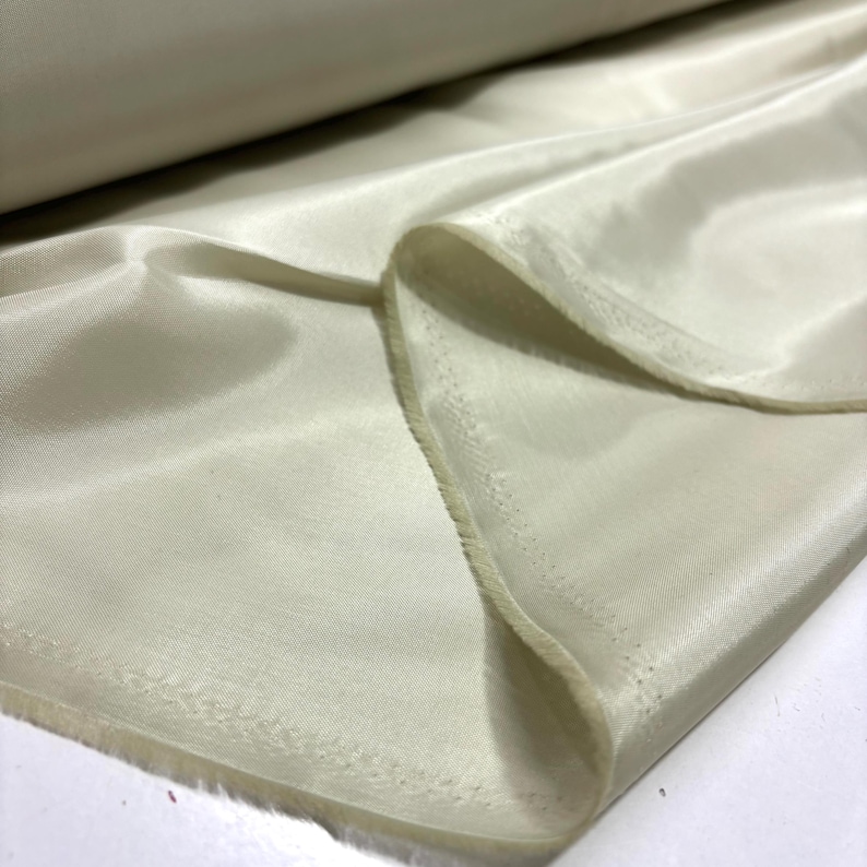 May include: A close-up of a roll of ivory-colored fabric. The fabric is smooth and shiny, with visible folds and a soft texture. The edges of the fabric are slightly frayed, suggesting a cut edge. The fabric appears to be lightweight and flowing.