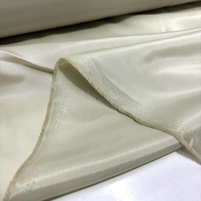 May include: Close-up of a roll of off-white fabric. The fabric has a smooth, silky texture and a slight sheen. The edge of the fabric is visible, showing a raw, unfinished edge. The fabric appears to be lightweight and suitable for various sewing projects.
