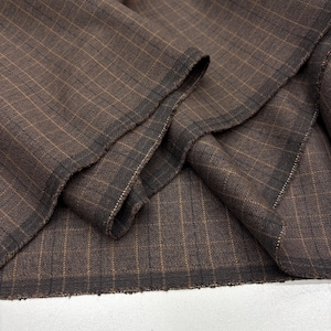 Può includere: Primo piano di tessuto marrone con motivo a quadri. Il tessuto ha un aspetto strutturato con linee marrone scuro e beige che formano una griglia. I bordi del tessuto sono sfilacciati e il materiale è piegato.
