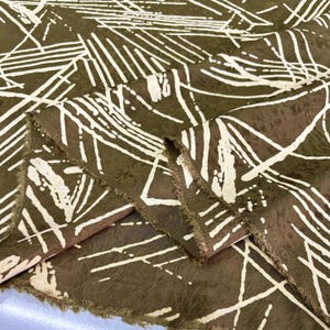 May include: Close-up of folded fabric with a dark olive-green background and a white, abstract linear pattern. The fabric has a textured appearance and frayed edges. The design is reminiscent of brushstrokes.