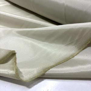 May include: Off-white satin fabric with a subtle sheen. The fabric is folded, revealing the texture and drape of the material. The edges are unfinished, showing the raw, natural fibers. The fabric is likely used for clothing, home decor, or craft projects.