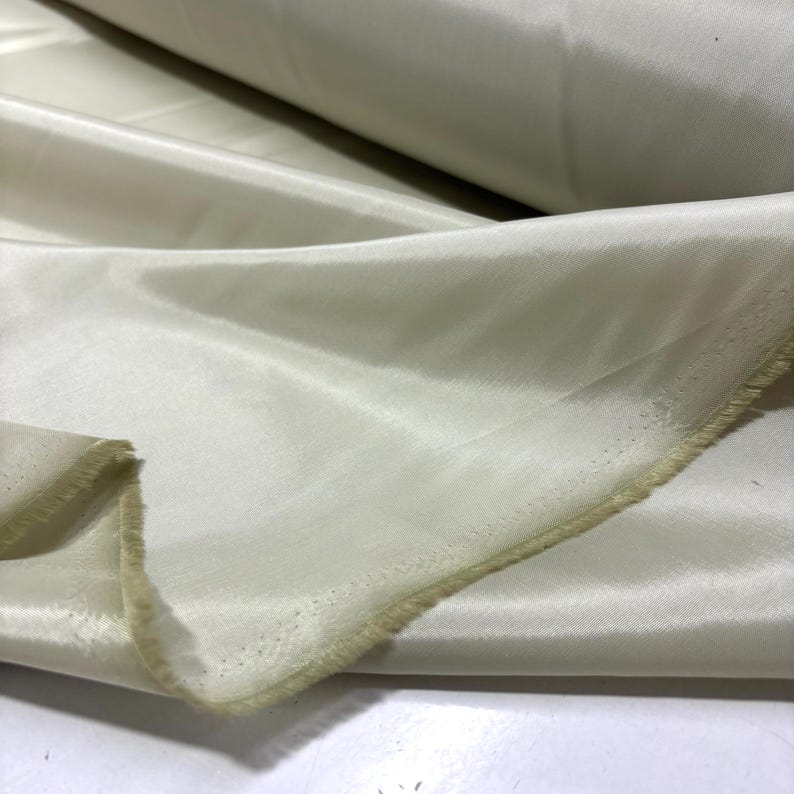 May include: Off-white satin fabric with a subtle sheen. The fabric is folded, revealing the texture and drape of the material. The edges are unfinished, showing the raw, natural fibers. The fabric is likely used for clothing, home decor, or craft projects.