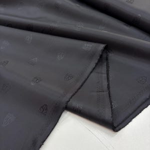 Puede incluir: Tela gris oscuro con un patrón sutil de diseños repetidos en forma de cresta. La tela está doblada, revelando la textura y el brillo del material. Los bordes muestran un corte en bruto sin terminar. Este textil es adecuado para ropa o decoración del hogar.