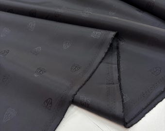 May include: Dark grey fabric with a repeating crest pattern. The fabric is folded, showing its texture and sheen. The edges are raw and unfinished. This textile is suitable for clothing or home furnishings. The fabric is likely measured in meters.