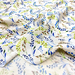 May include: White fabric with a floral pattern featuring blue, green, and brown botanical elements. The fabric is folded, showing the detailed design of the flowers and leaves. The overall aesthetic is delicate and nature-inspired.