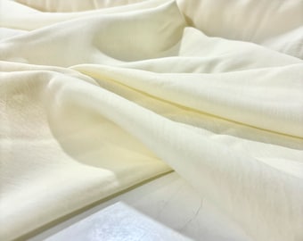Puede incluir: Primer plano de una tela lisa de color blanco roto, con una textura suave y caída. La tela está doblada y dispuesta de forma que se muestra su textura y caída.