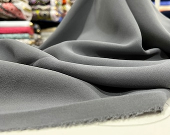 May include: A close-up of a soft, gray fabric with a slightly textured surface. The fabric is draped and folded, showcasing its flowy and drapable nature.