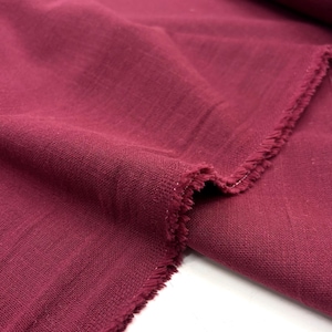Könnte beinhalten: Nahaufnahme eines bordeauxfarbenen Stoffes mit strukturiertem Gewebe. Der Stoff ist gefaltet und zeigt eine rohe, ausgefranste Kante. Die Farbe ist ein tiefer, satter Rotton mit leichtem Glanz, was auf ein Textil aus Naturfasern hindeutet.
