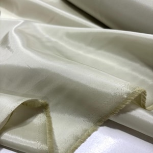 May include: Close-up of a roll of ivory-colored fabric. The material has a smooth, shiny surface with visible folds and a slightly frayed edge. The fabric appears to be lightweight and suitable for clothing or decorative projects.