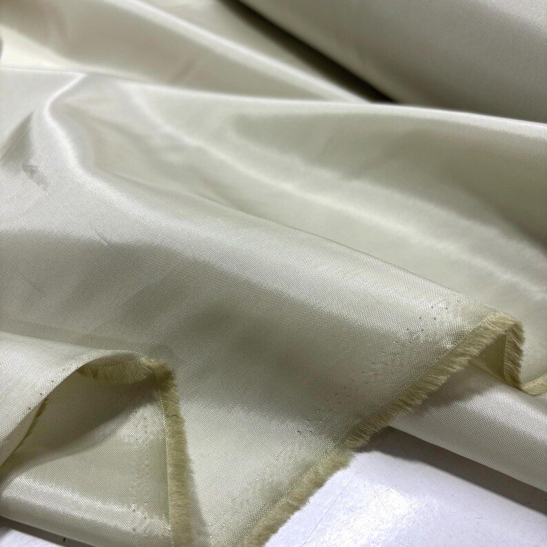May include: Close-up of a roll of ivory-colored fabric. The material has a smooth, shiny surface with visible folds and a slightly frayed edge. The fabric appears to be lightweight and suitable for clothing or decorative projects.