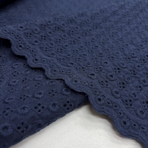 Op de afbeelding: Close-up van marineblauwe broderie anglaise stof. De stof heeft een geschulpte rand en een herhalend patroon van kleine, geborduurde bloemmotieven. De stof is gevouwen en toont de textuur en diepte van het materiaal.