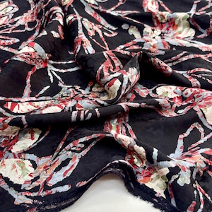 May include: Black fabric with a floral pattern in red, white, and light blue. The fabric is folded, showing the texture and the frayed edge. The design has a vintage or botanical feel.
