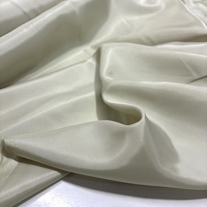 May include: Ivory-colored fabric with a silky sheen. The material is draped, creating soft folds and shadows. The fabric appears lightweight and flowing, suggesting it could be used for apparel or decorative purposes.