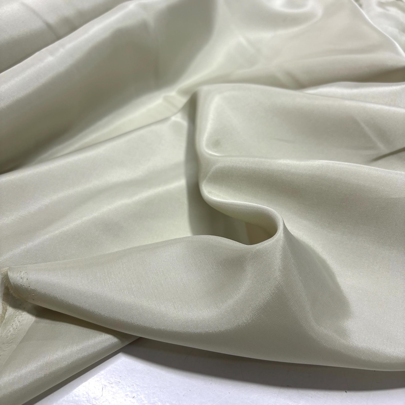 May include: Ivory-colored fabric with a silky sheen. The material is draped, creating soft folds and shadows. The fabric appears lightweight and flowing, suggesting it could be used for apparel or decorative purposes.