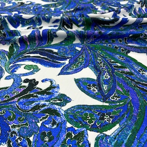 Puede incluir: Primer plano de una tela blanca con un vibrante estampado de cachemira. El diseño presenta grandes motivos florales y de hojas estilizados en tonos de azul, verde y morado. La tela tiene un aspecto suave y fluido.