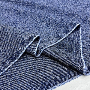 May include: Close-up of a piece of blue denim fabric. The fabric has a textured appearance with a diagonal weave pattern. The edges of the fabric are frayed, showing the raw edge of the material.