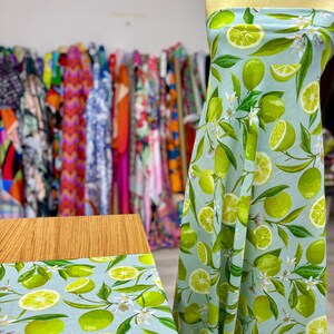 May include: Light blue fabric with a vibrant lime and floral pattern. The design features whole and sliced limes, green leaves, and white blossoms. The fabric is draped over a form and also displayed flat on a wooden surface, showcasing the repeating pattern.