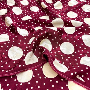 May include: Burgundy fabric with a white polka dot pattern. The fabric is folded, revealing its texture and sheen. The polka dots are of varying sizes, creating a dynamic design. Ideal for dressmaking or crafting.