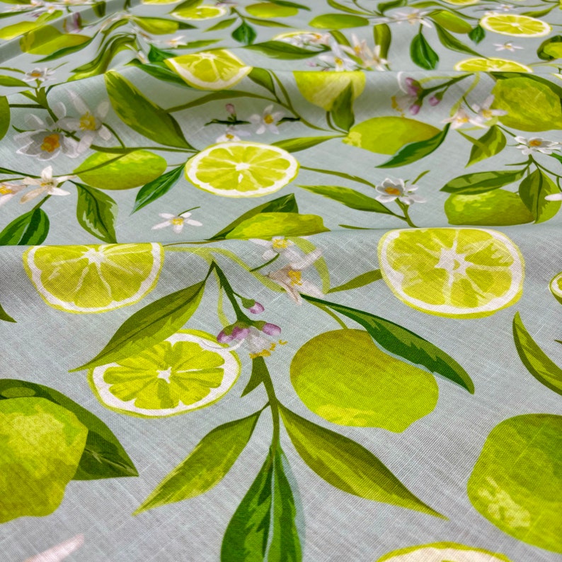 May include: Fabric with a light blue background featuring a pattern of green limes, lime slices, green leaves, and white blossoms. The limes are whole and sliced, with a watercolor style. The fabric is suitable for various sewing projects.