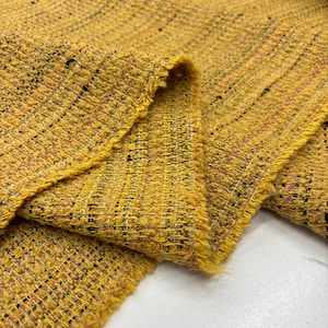 Op de afbeelding: Close-up van een textiel met een levendige gele tint. De weefselstructuur is zichtbaar, met een mix van geel, zwart en vlekken van andere kleuren. De randen van de stof zijn gerafeld, wat bijdraagt aan de rustieke charme. Dit textiel is geschikt voor diverse knutselprojecten.