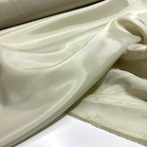 May include: Close-up of a cream-colored fabric with a silky, flowing texture. The material is draped, showing folds and highlights. The fabric appears to be a lightweight material, possibly for apparel or home decor.