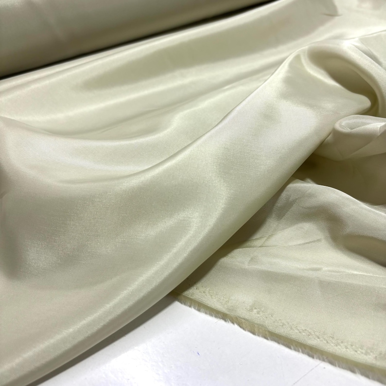 May include: Close-up of a cream-colored fabric with a silky, flowing texture. The material is draped, showing folds and highlights. The fabric appears to be a lightweight material, possibly for apparel or home decor.
