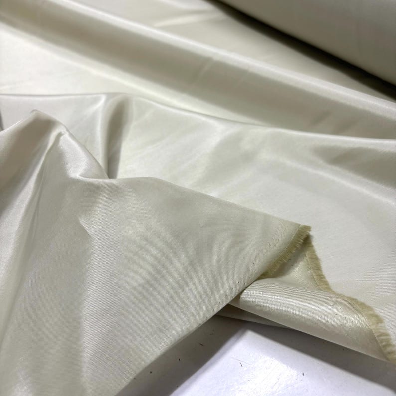 May include: Off-white fabric with a silky sheen. The fabric is draped, showing folds and texture. The material appears to be lightweight and flowing, with a subtle luster. The fabric is likely used for apparel or home decor.