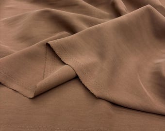 Op de afbeelding: Close-up van een opgevouwen stuk stof in een warme, aardse bruine kleur. De stof heeft een zachte, vloeiende textuur met zichtbare vouwen en een rafelige rand. Het materiaal lijkt lichtgewicht en geschikt voor kleding of knutselprojecten.