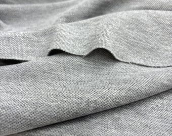 Op de afbeelding: Close-up van grijze stof met een getextureerd, wafelachtig patroon. De stof is gevouwen en gedrapeerd, wat de textuur- en kleurvariaties laat zien. Het materiaal lijkt een gebreide of geweven textiel te zijn, geschikt voor kleding of woondecoratie.