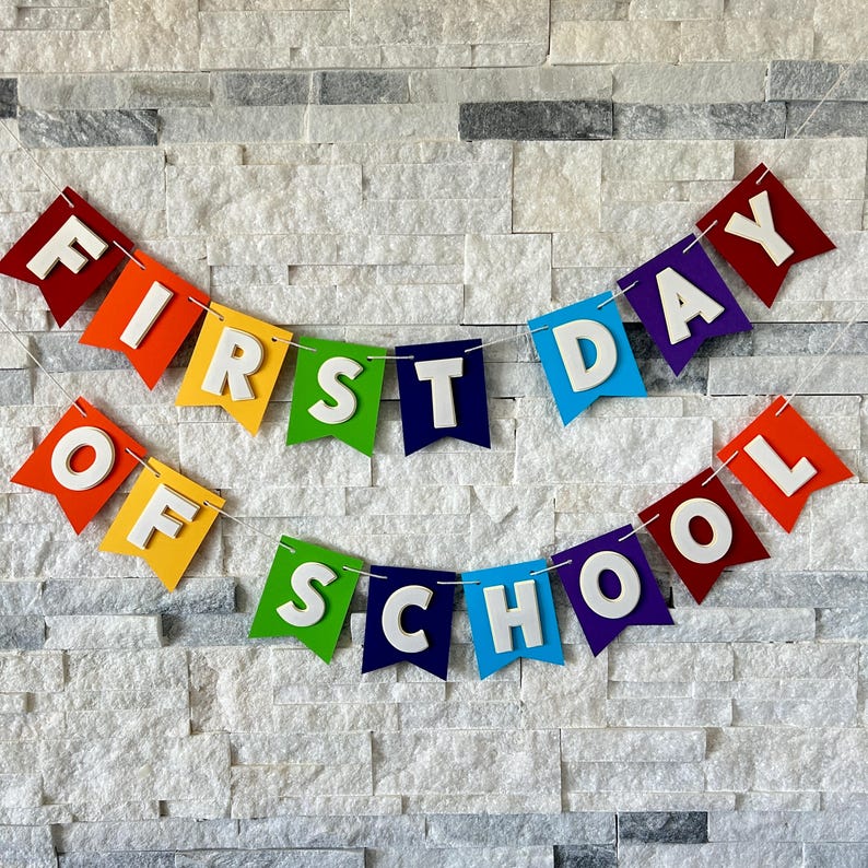 Puede incluir: Una colorida pancarta con las palabras "FIRST DAY OF SCHOOL" en letras blancas. La pancarta est&aacute; hecha de banderas de fieltro en colores del arco&iacute;ris, unidas con una fina cuerda blanca, y se muestra contra una pared de piedra blanca texturizada.