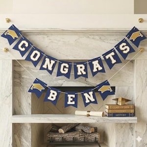 May include: A navy blue and gold graduation banner that reads "CONGRATS BEN". The banner is strung across a fireplace mantel. A rolled diploma, books, and a graduation cap are also on the mantel.