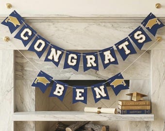 May include: A navy blue and gold graduation banner reading "CONGRATS BEN" strung across a fireplace mantel. A rolled diploma, books, and a graduation cap are also on the mantelpiece.