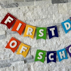 Puede incluir: Una colorida guirnalda con el texto "FIRST DAY OF SCHOOL". Las banderas son rojas, naranjas, amarillas, verdes, azules y moradas, con letras blancas en 3D. El fondo es una pared de piedra gris claro texturizada. Perfecto para el regreso a clases.