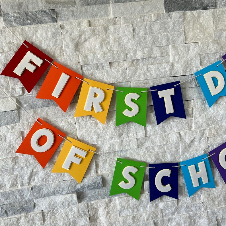 Puede incluir: Una colorida guirnalda con el texto "FIRST DAY OF SCHOOL". Las banderas son rojas, naranjas, amarillas, verdes, azules y moradas, con letras blancas en 3D. El fondo es una pared de piedra gris claro texturizada. Perfecto para el regreso a clases.