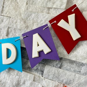 Puede incluir: Una colorida pancarta que deletrea "DAY" con banderas individuales en azul, morado y rojo. Cada bandera tiene una letra blanca tridimensional adjunta. La pancarta est&aacute; ensartada con una cuerda blanca sobre un fondo gris claro texturizado.