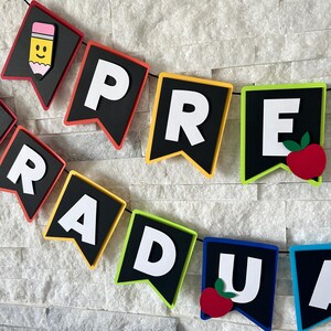 K&ouml;nnte beinhalten: Ein schwarz-wei&szlig;es Banner mit den Worten "PRE-GRAD" in wei&szlig;en Buchstaben. Das Banner ist mit bunten R&auml;ndern und roten Apfel-Ausschnitten verziert.