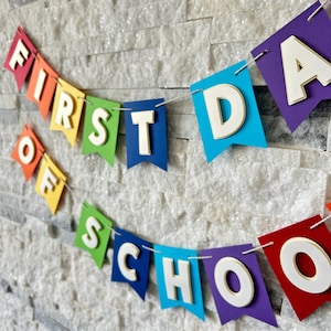 Puede incluir: Una colorida pancarta con las palabras "FIRST DAY OF SCHOOL" en letras blancas con bordes dorados. Las banderas de la pancarta son de colores del arco&iacute;ris y est&aacute;n ensartadas en una cuerda blanca, perfecta para una celebraci&oacute;n de regreso a clases.