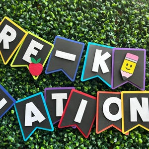 K&ouml;nnte beinhalten: Ein farbenfrohes Banner mit den Worten "Pre-K Graduation" in wei&szlig;en Buchstaben auf schwarzen Flaggen. Jede Flagge hat einen andersfarbigen Rand. Die Flaggen sind mit einem roten Apfel und einem gelben Bleistift mit einem Smiley verziert.