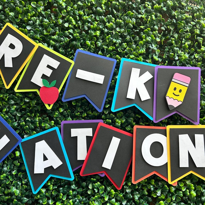 K&ouml;nnte beinhalten: Ein farbenfrohes Banner mit den Worten "Pre-K Graduation" in wei&szlig;en Buchstaben auf schwarzen Flaggen. Jede Flagge hat einen andersfarbigen Rand. Die Flaggen sind mit einem roten Apfel und einem gelben Bleistift mit einem Smiley verziert.