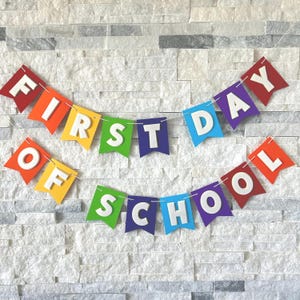 Puede incluir: Una colorida pancarta con las palabras "FIRST DAY OF SCHOOL" en letras blancas. Cada palabra est&aacute; en una bandera de bander&iacute;n de colores del arco&iacute;ris, unidas por una cuerda. El fondo es una pared de piedra gris claro.
